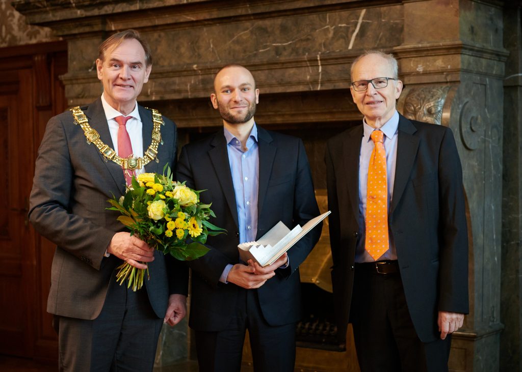 Drei Männer, der linke trägt eine Bürgermeisterkette, der mittlere eine Urkunde, der rechte eine leuchtend-orange Krawatte.