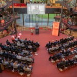 Blick von oben ins Foyer der HSU/UniBw H. Die Leitung der HSU/UniBw H und Ehrengäste sitzen in Stuhlreihen und lauschen dem Redner am Rednerpult.